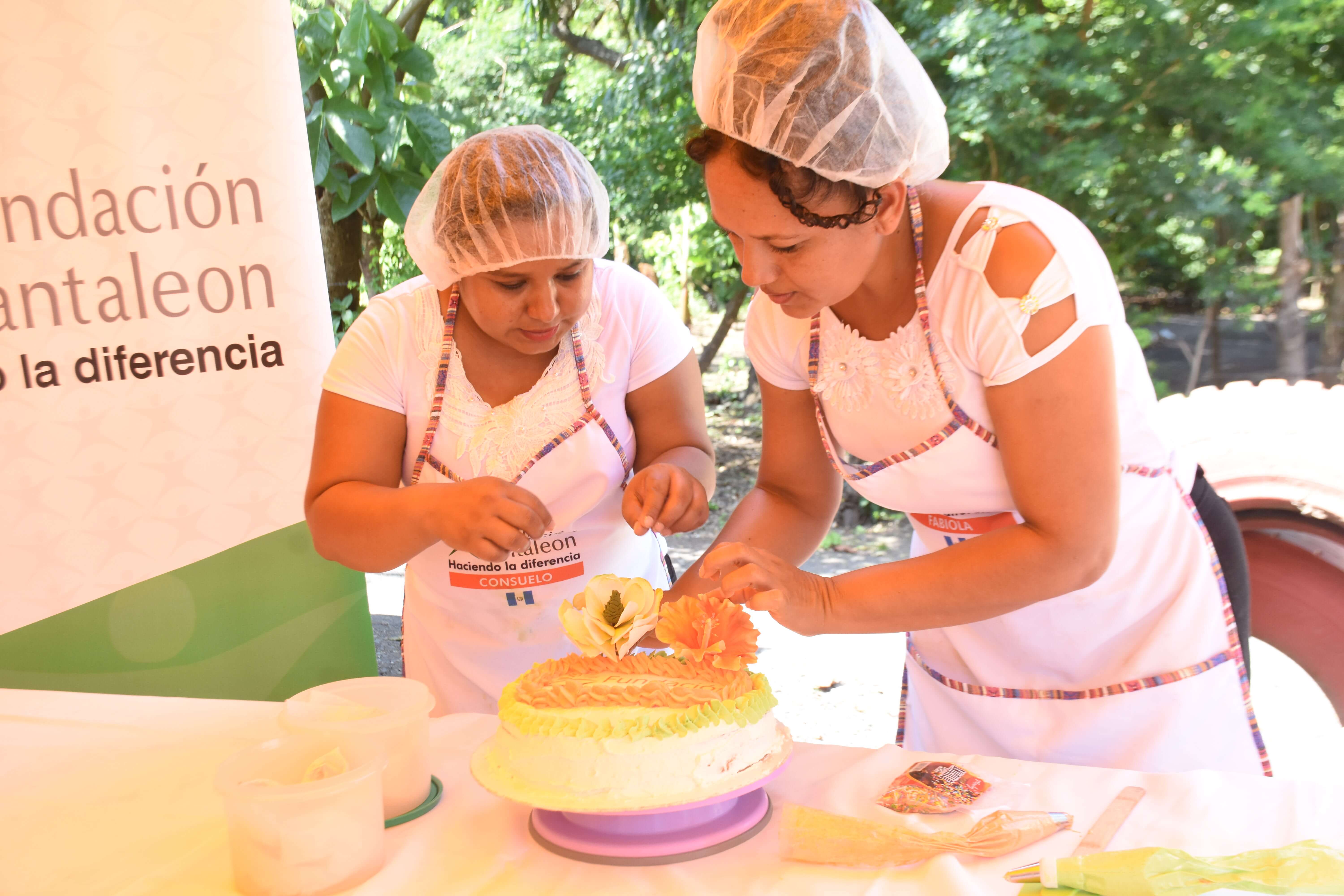 Dos mujeres con cofias y delantales blancos decoran un pastel con flores de azúcar en una mesa al aire libre, con un cartel de Fundación Pantaleon al fondo.