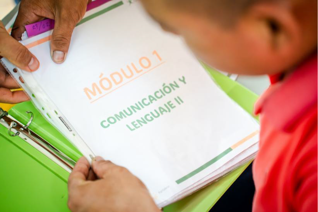 Una persona sostiene un cuadernillo con la portada que dice 'Módulo 1 Comunicación y Lenguaje II' mientras otra persona observa, sobre una mesa verde.