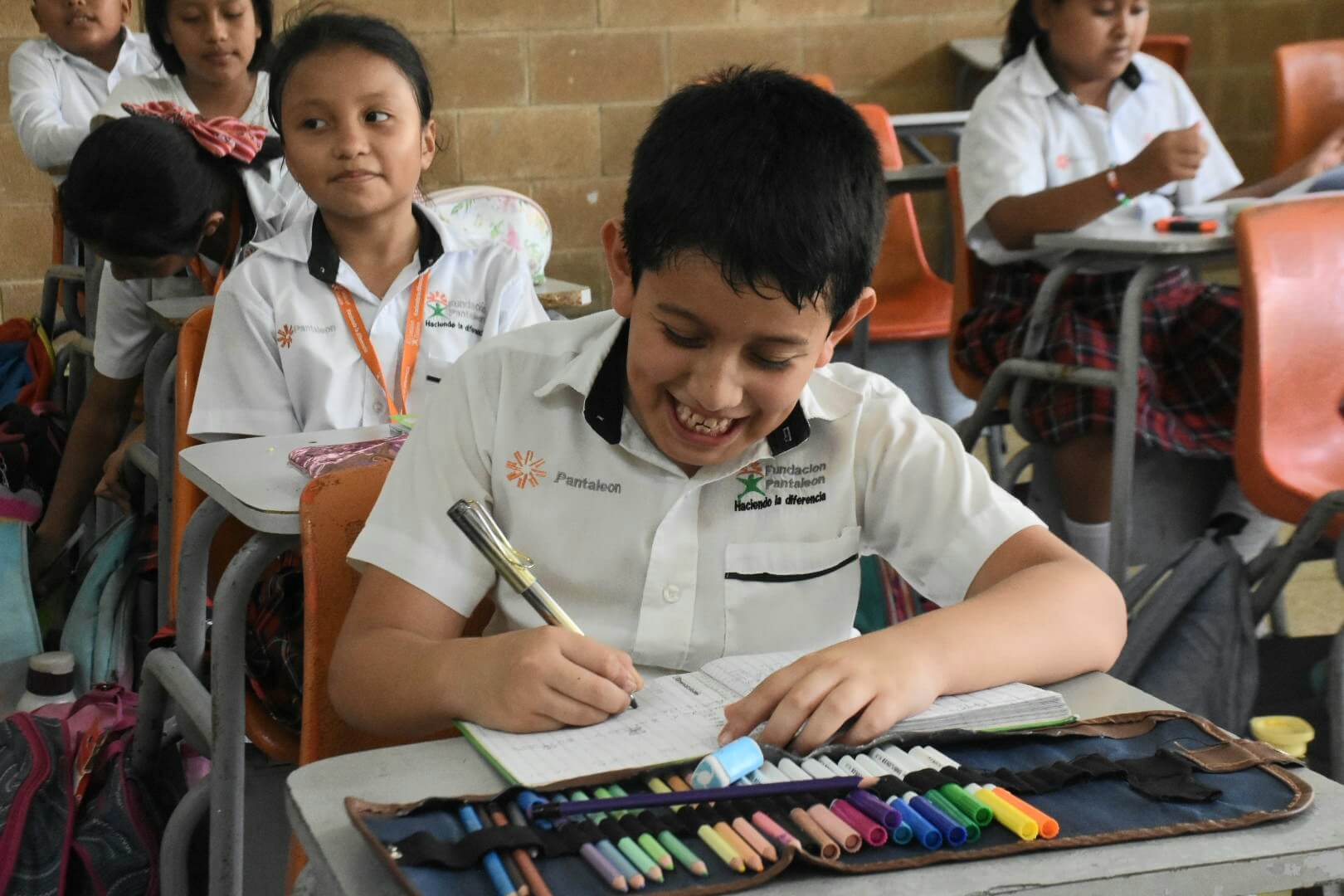 Niño sonriente escribiendo en un cuaderno en su escritorio de salón, con crayones y marcadores organizados al frente, rodeado de otros niños en uniforme escolar blanco.