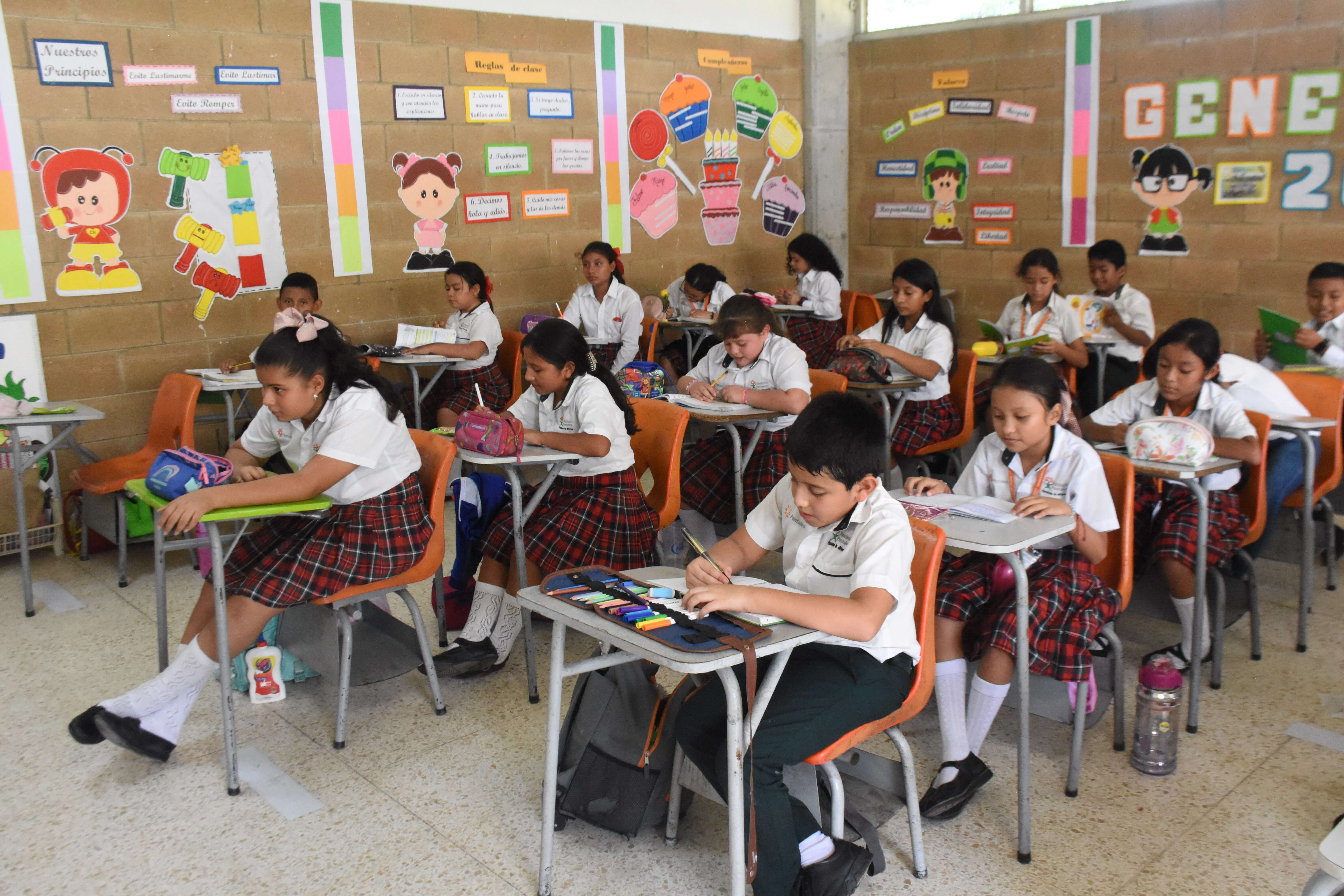 Estudiantes vestidos con uniformes escolares sentados en escritorios en un salón de clases con paredes decoradas con carteles coloridos y dibujos animados.
