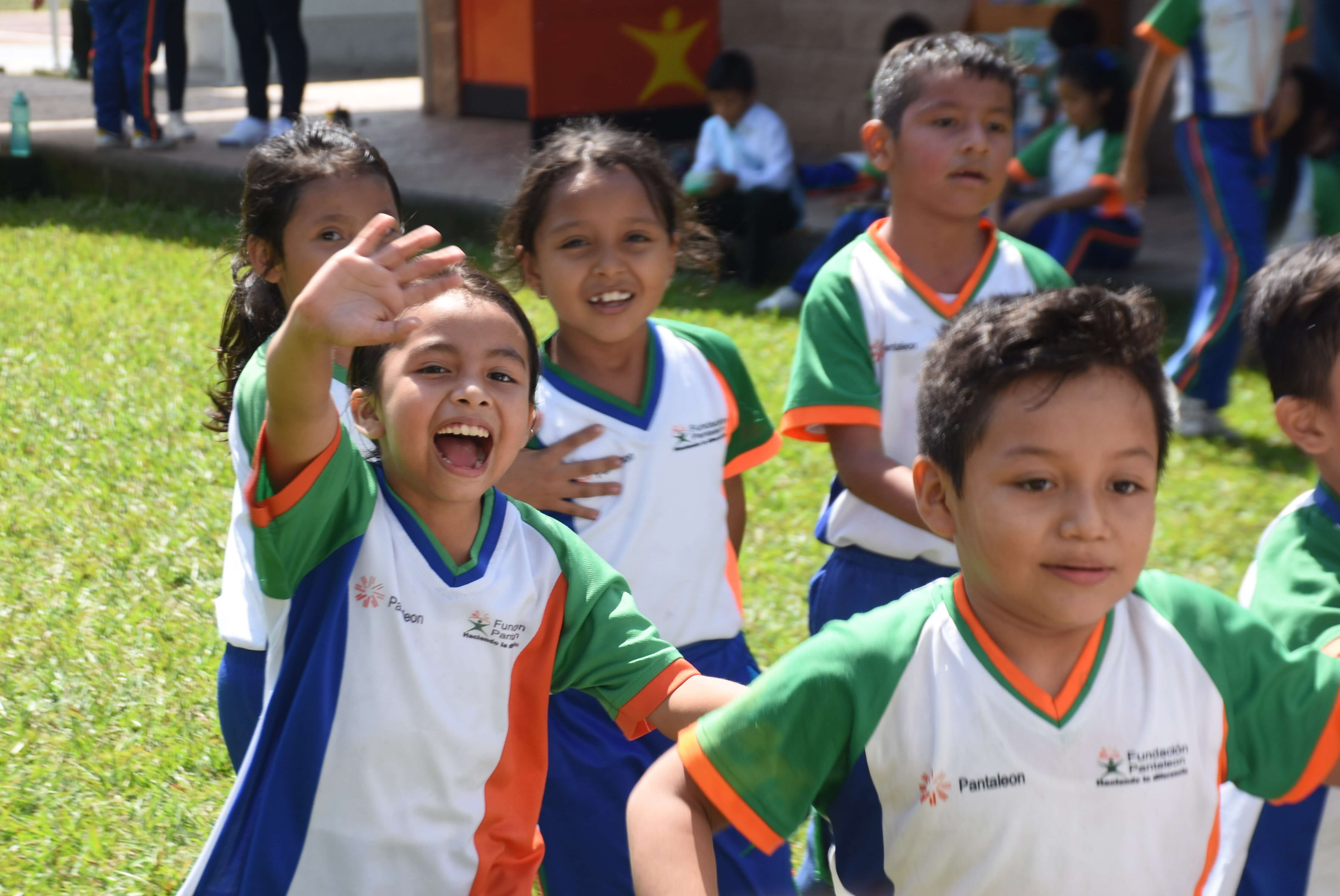 Niños con uniformes deportivos blancos con verde, azul y naranja jugando y sonriendo al aire libre en un área césped, uno de ellos saluda con la mano levantada y la boca abierta en señal de alegría.