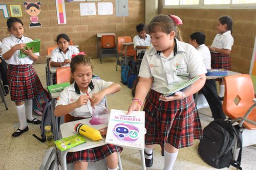 Niñas en uniforme escolar con camisa blanca y falda de cuadros rojos en un salón de clases con paredes de bloques. Una niña está sentada en un pupitre manipulando un objeto transparente con líquido rosado, mientras otra niña le ofrece un cuaderno de actividades. Más estudiantes están sentados o parados en el fondo, algunos con cuadernos y mochilas.