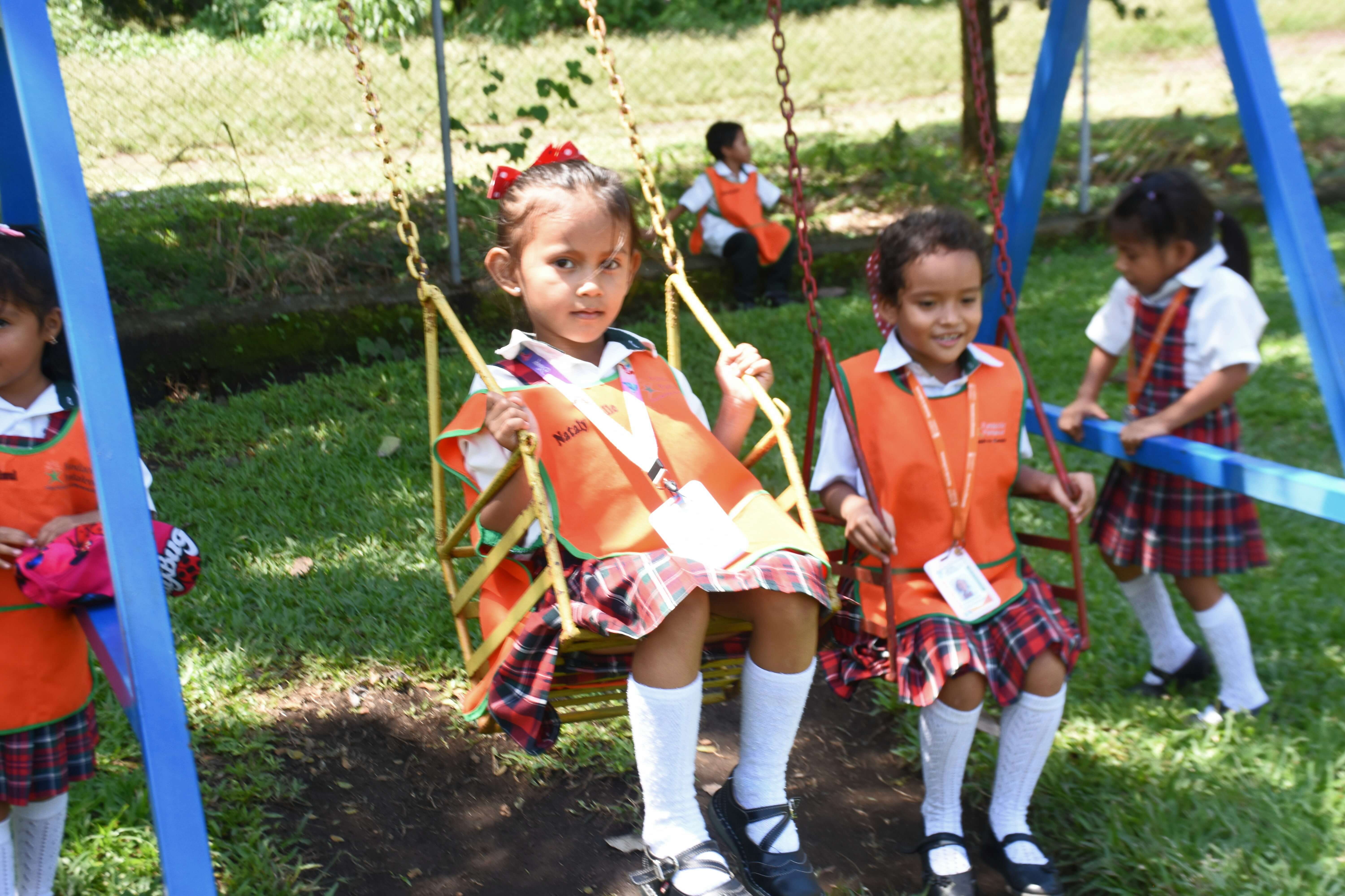 Niñas uniformadas con faldas a cuadros y chalecos naranjas jugando en columpios en un área verde al aire libre.