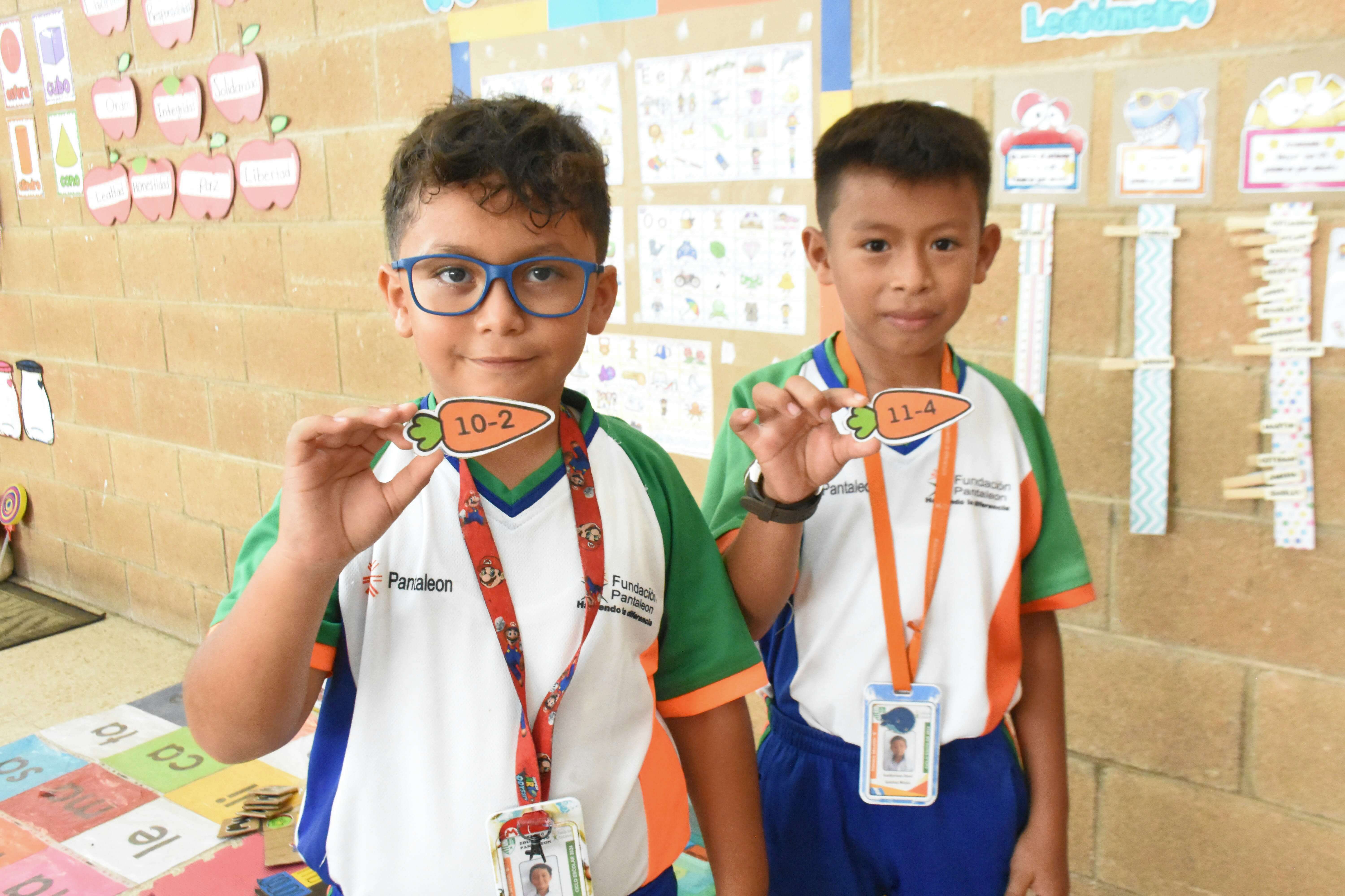 Dos niños en uniforme escolar sostienen recortes en forma de zanahoria con restas escritas: 10-2 y 11-4, dentro de un aula con decoraciones educativas en la pared.