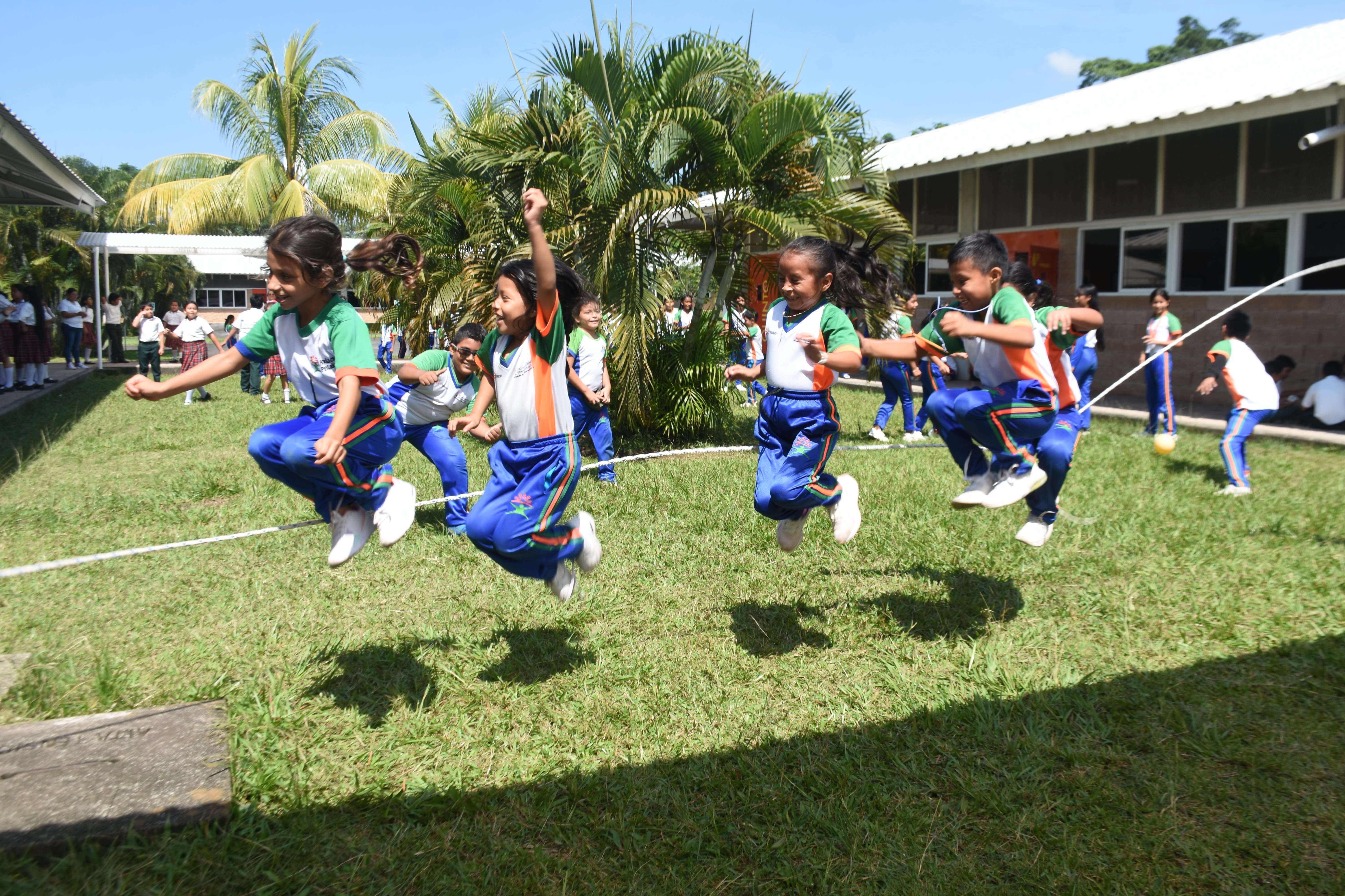 Niños y niñas en uniforme escolar saltando en grupo la cuerda larga en un patio con pasto y palmeras, mientras otros niños observan y juegan alrededor bajo un cielo azul despejado.