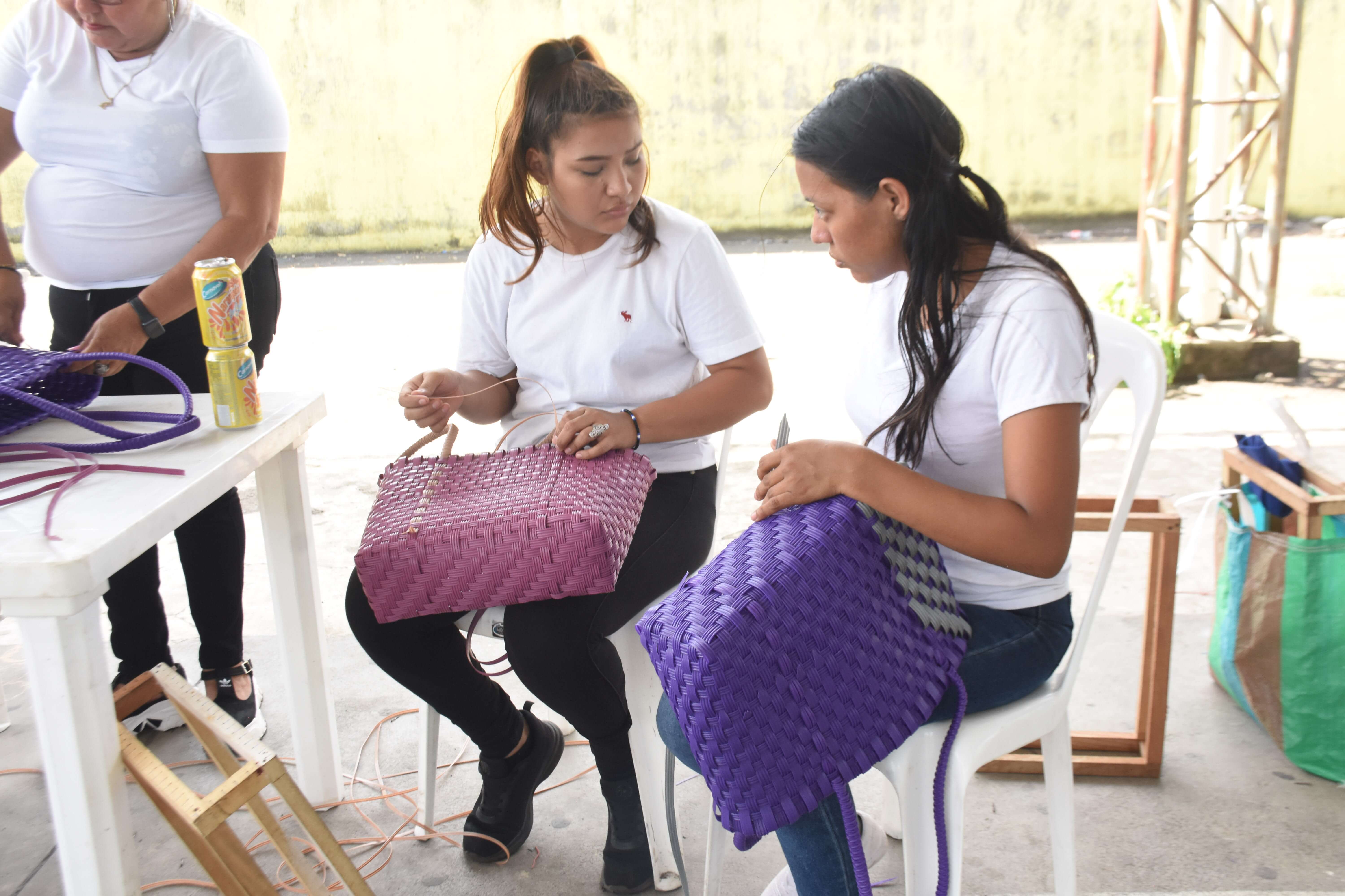 Dos mujeres sentadas en sillas blancas tejiendo bolsas de plástico en un espacio abierto, una con bolsa morada y la otra con bolsa lila, mientras otra persona de pie trabaja en una mesa blanca con latas de bebida encima.