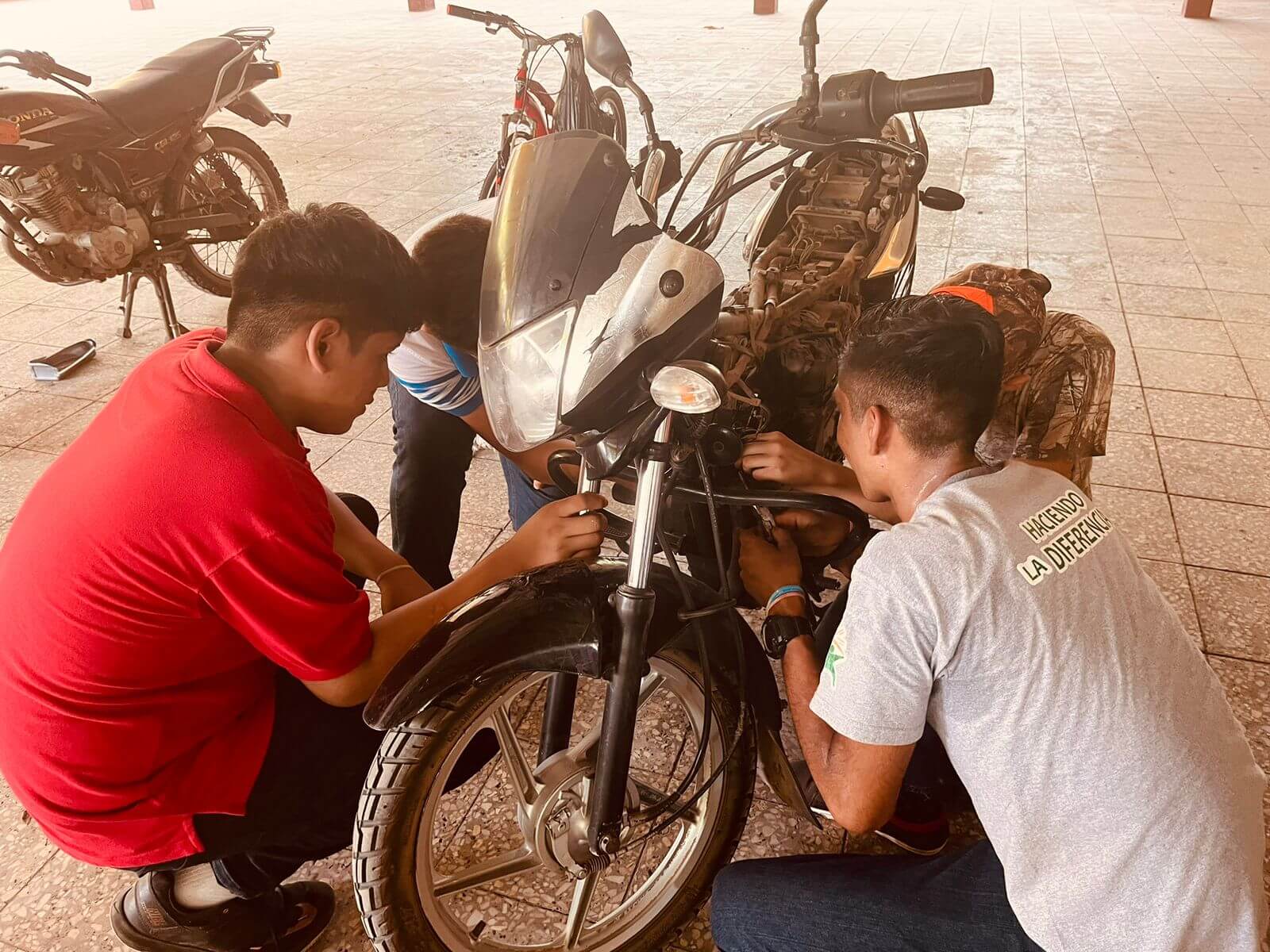 Cuatro jóvenes están reparando una motocicleta negra sin asiento en un piso de baldosas, uno lleva camisa roja, otro gris con el texto "Haciendo la diferencia", y otro camuflado.