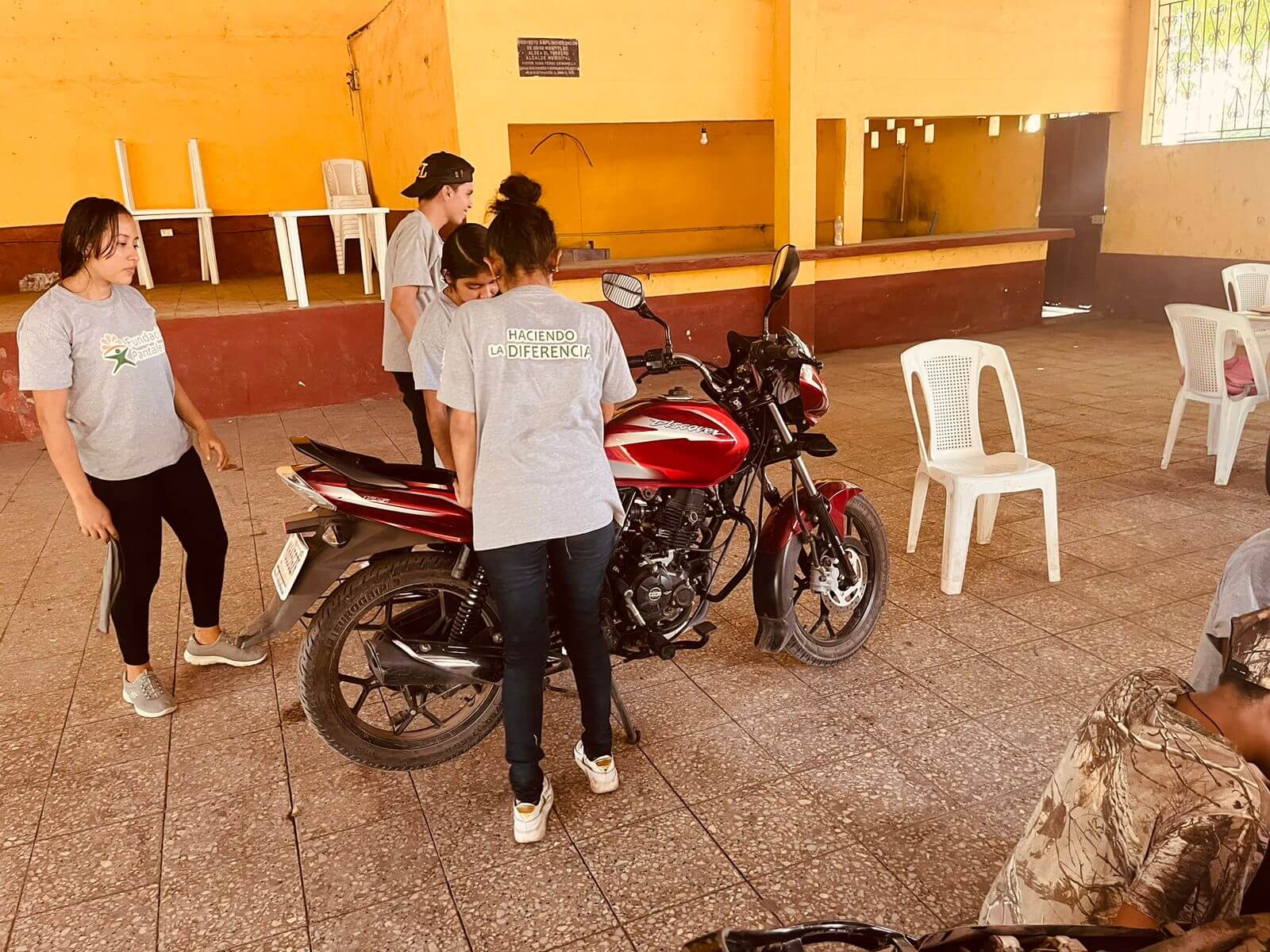 Un grupo de jóvenes con camisetas grises están reunidos alrededor de una motocicleta roja dentro de un salón con paredes amarillas y sillas plásticas blancas.