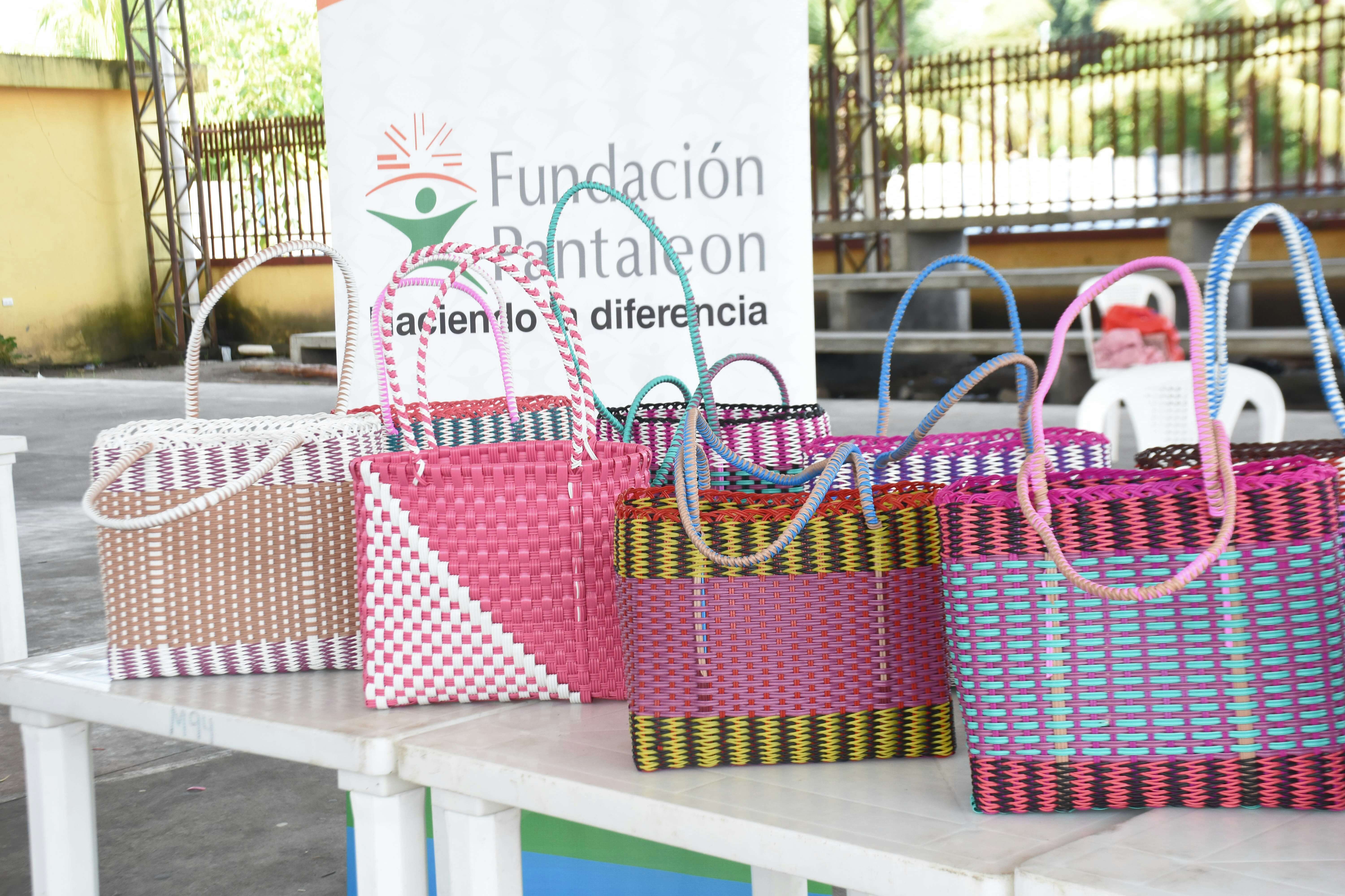 Bolsas tejidas con colores variados en una mesa blanca en un entorno al aire libre, al fondo un cartel que dice Fundación Pantaleon haciendo la diferencia.
