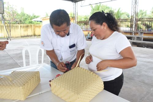 Hombre y mujer trabajando juntos en la elaboración de una bolsa tejida de palma en una mesa blanca, en un espacio techado al aire libre.