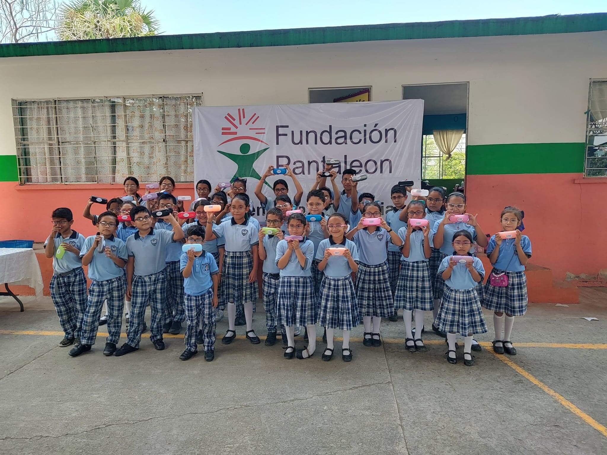 Grupo de niños y niñas en uniforme escolar azul y falda o pantalón a cuadros posando frente a una pancarta que dice Fundación Pantaleon, cada uno sosteniendo una caja colorida en las manos.