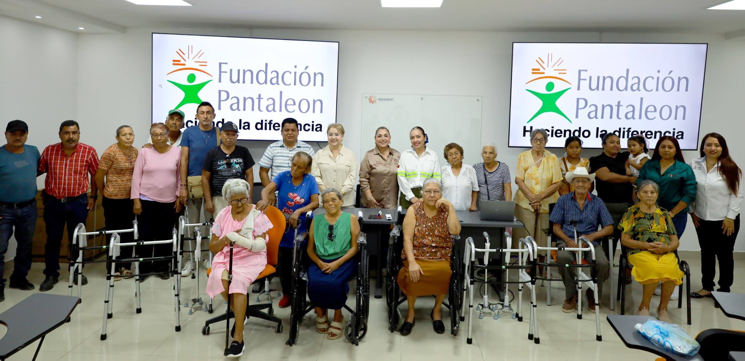 Grupo diverso de personas, incluyendo adultos mayores, algunos en sillas de ruedas y otros con andadores, posando en una sala con dos pantallas que muestran el logo y nombre "Fundación Pantaleón Haciendo la diferencia".