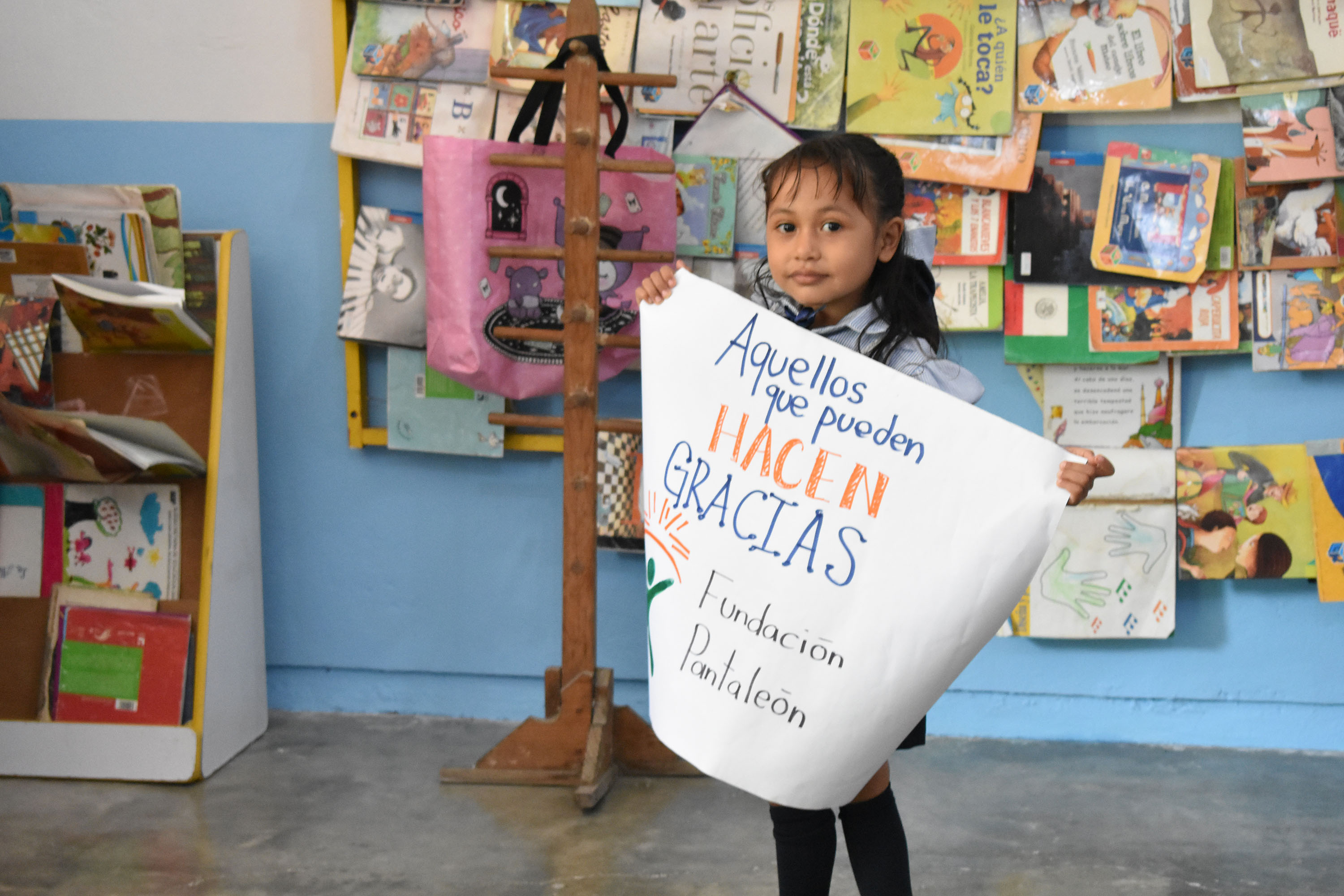 Niña en uniforme escolar sosteniendo un cartel que dice 'Aquellos que pueden HACEN GRACIAS Fundación Pantaleón' en un ambiente con estantes y libros detrás.