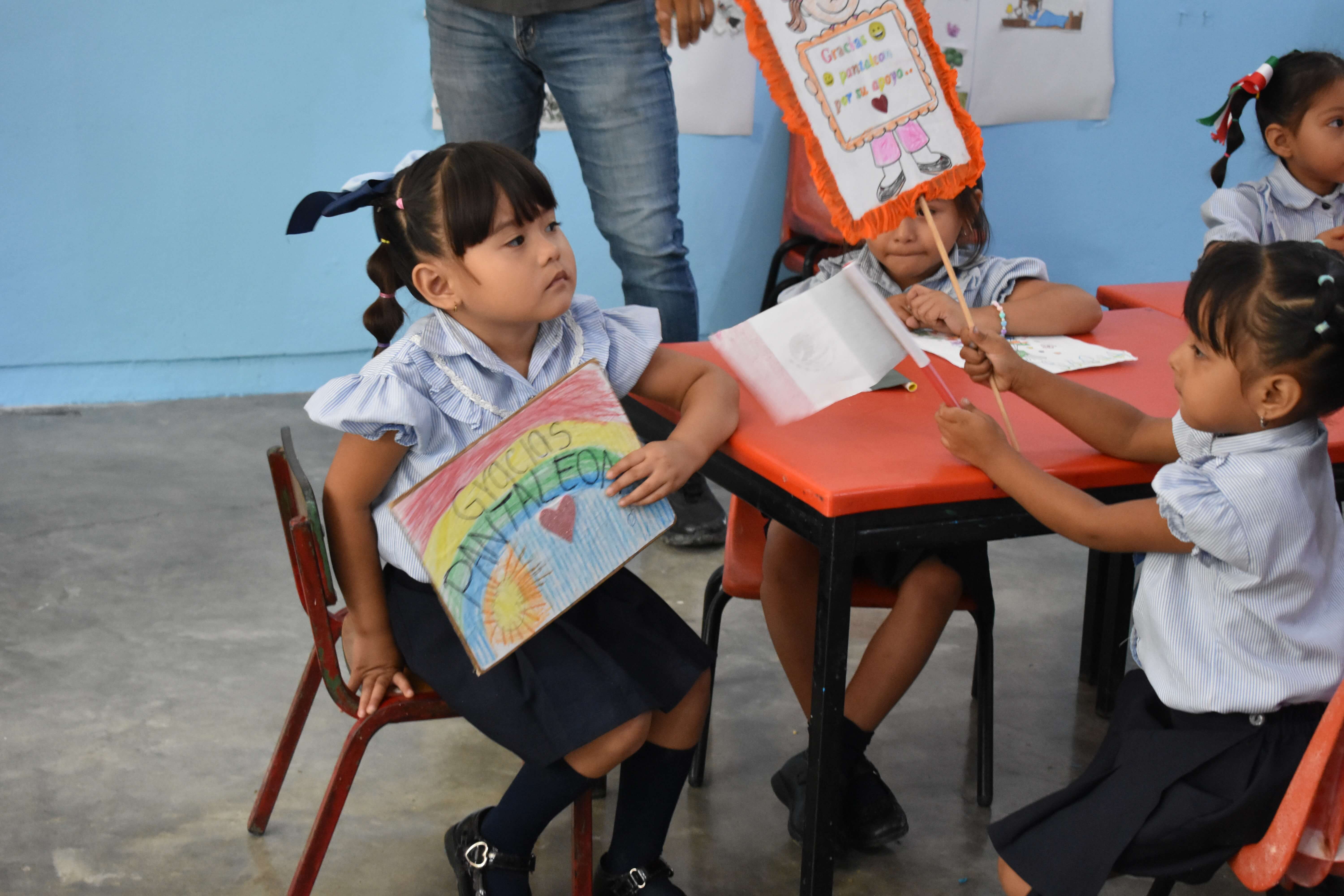 Niñas en uniforme escolar sentadas en sillas rojas y una mesa naranja dentro de un salón con paredes azules, una sostiene un cartel que dice ' Gracias DANANACLEON ' con un dibujo de un sol y colores, otra sostiene una bandera blanca y un cartel con un dibujo y las palabras 'Gracias a pandadero por su apoyo'.