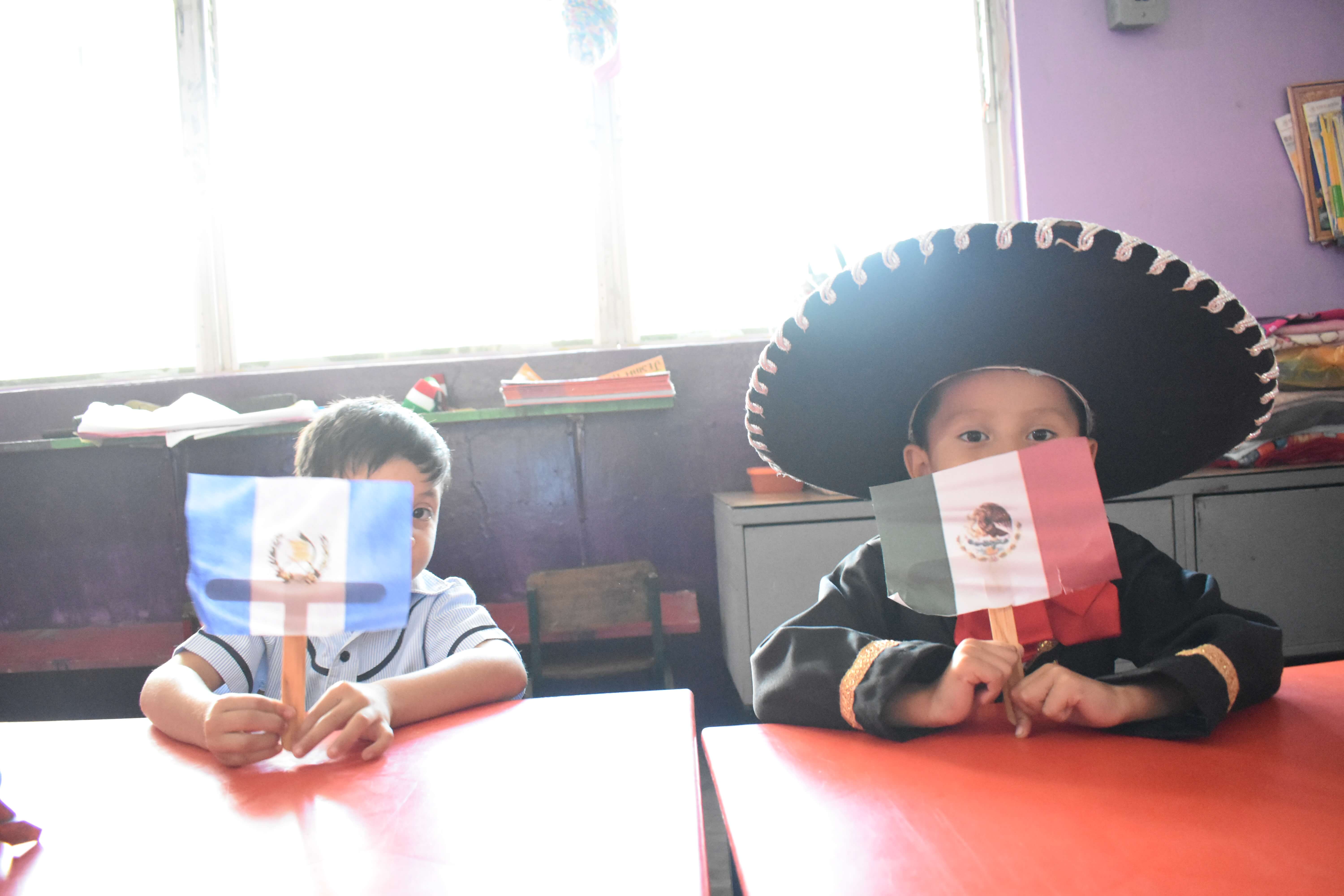 Dos niños sentados en mesas rojas sostienen banderas pequeñas; el niño a la izquierda tiene la bandera de Guatemala, y el niño a la derecha, que lleva un sombrero tradicional mexicano, sostiene la bandera de México.