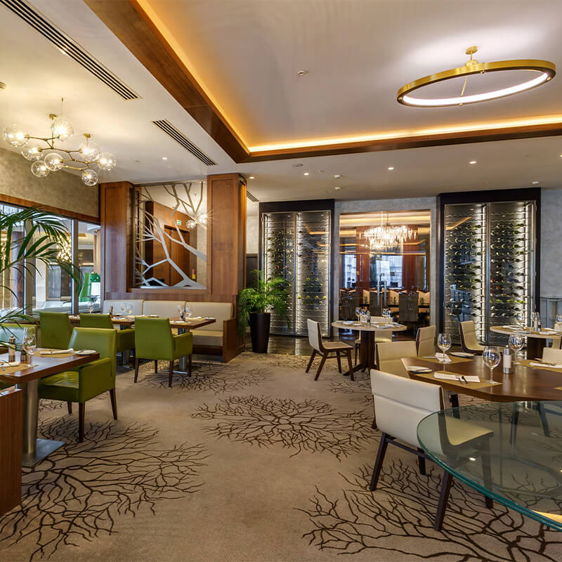 Modern upscale restaurant interior with green and cream chairs, round tables, decorative carpet, and illuminated wine displays.