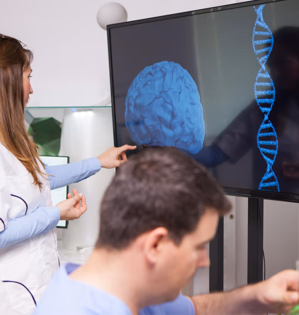 Woman in a white coat pointing to a 3D blue brain and DNA strand on a large monitor while a man works in the foreground.