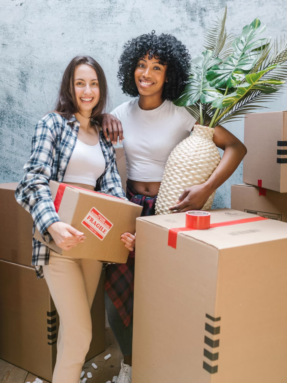 Twee lachende vrouwen omringd door verhuisdozen, één draagt een doos met een fragiel-label en de ander houdt een grote vaas met groen blad.