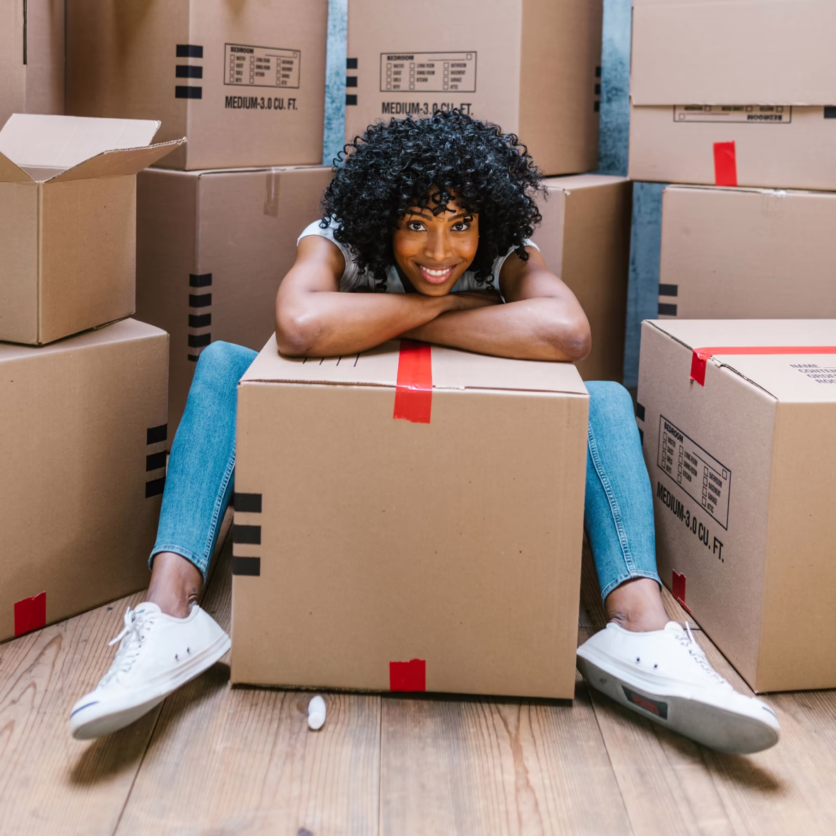 Vrouw zit op houten vloer tussen dozen en leunt glimlachend op een doos met rode tape.