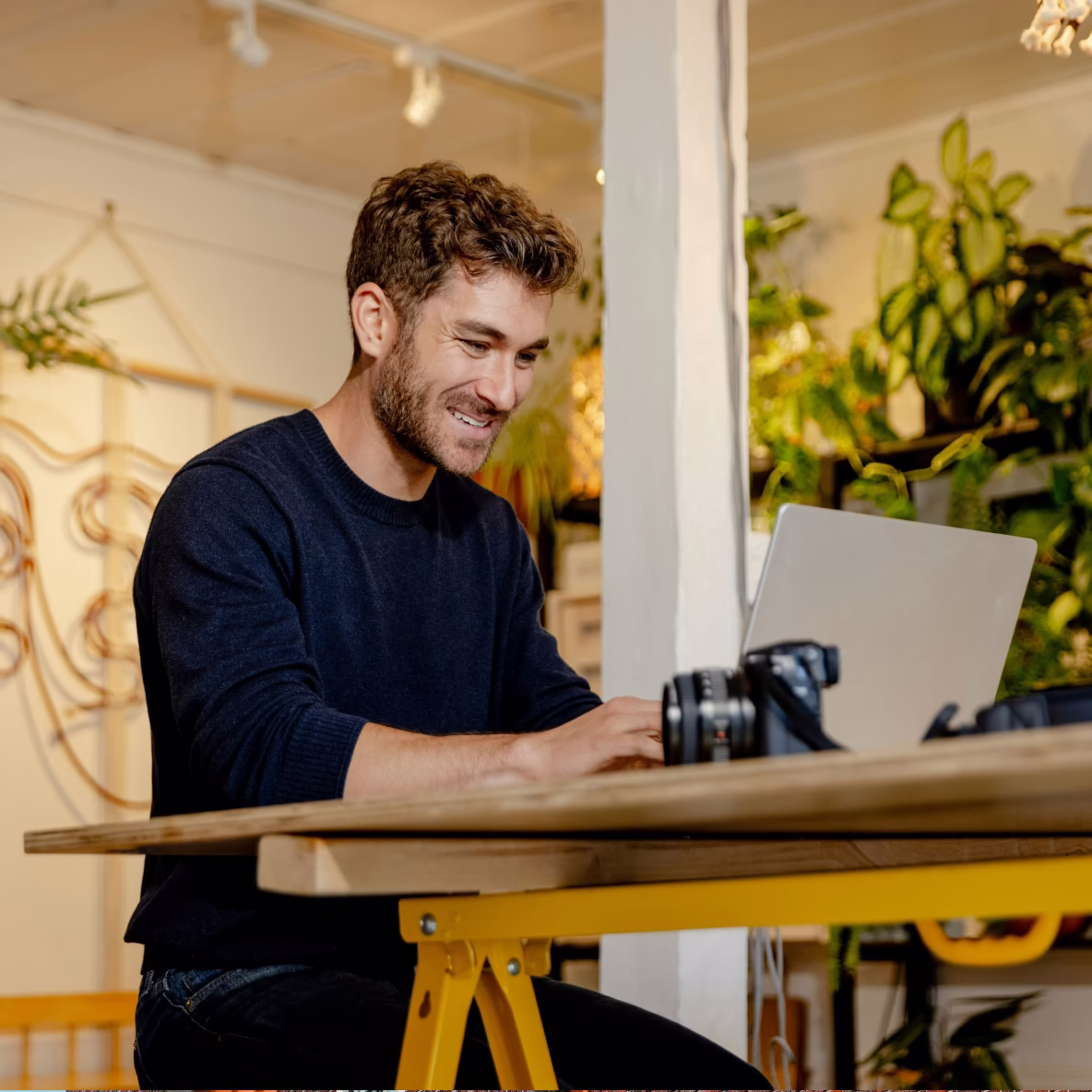Jonge man die glimlachend op een laptop werkt aan een houten tafel met een camera erop in een gezellige, groene binnenruimte.