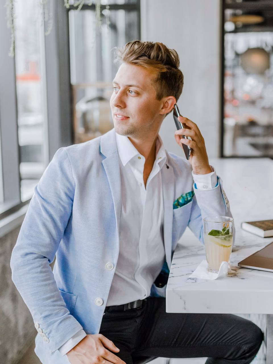 Jonge man in lichtblauw colbert en witte blouse zit aan een marmeren tafel en belt op zijn mobiele telefoon terwijl hij uit het raam kijkt.