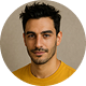 Portrait of a young man with short dark hair, a trimmed beard, and a yellow shirt against a beige background.