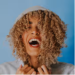 Person with curly hair partially covering their face, laughing, wearing a light-colored hoodie against a blue background.