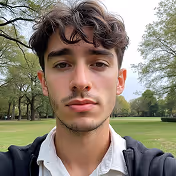 Young man with curly dark hair and a light beard wearing a white shirt and black jacket in a park with green trees and grass.