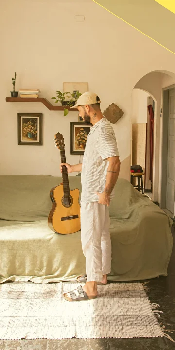 Man wearing a beige cap and striped shirt holding an acoustic guitar standing next to a green-covered sofa in a cozy room.