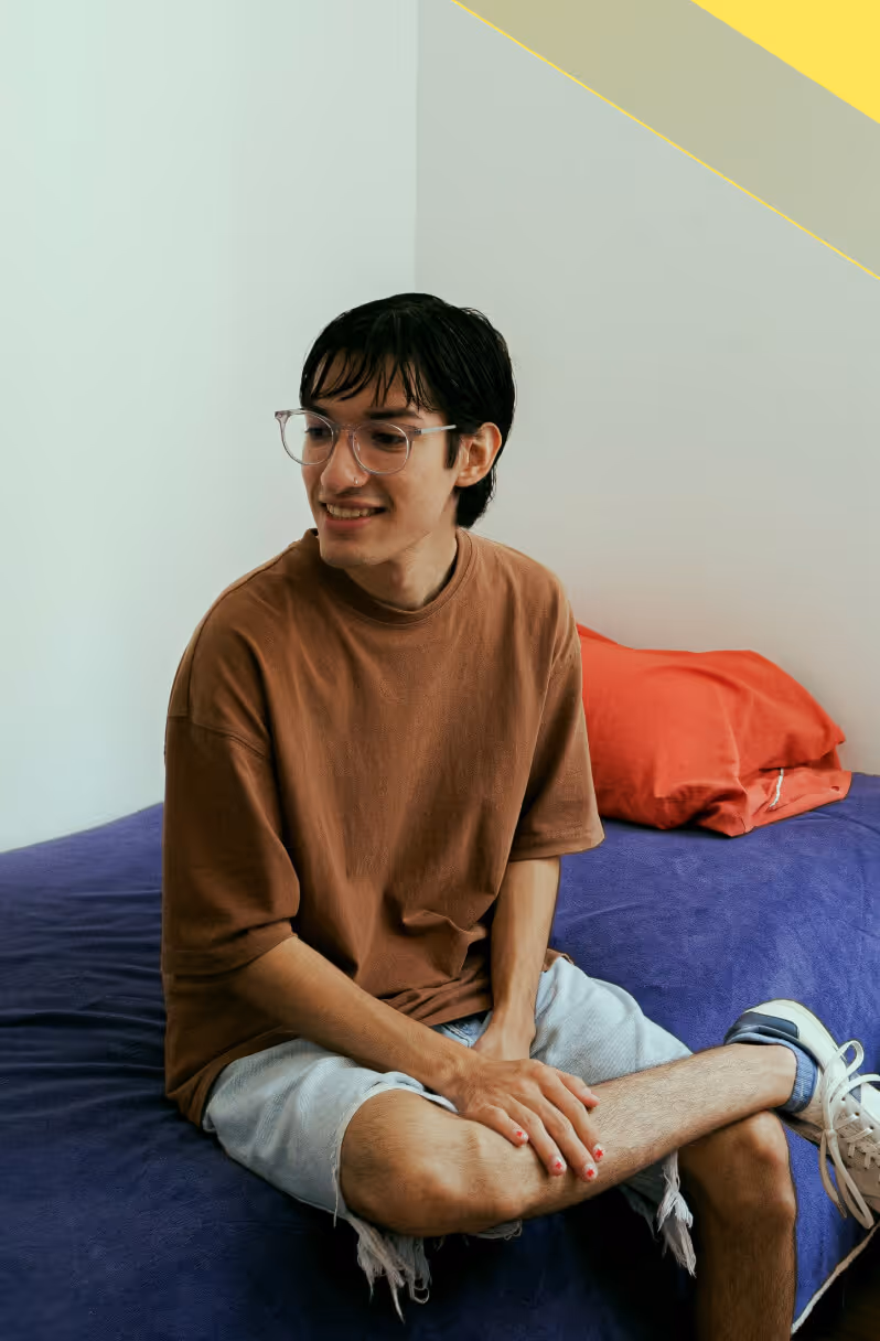Young person with short dark hair and glasses sitting cross-legged on a bed with a blue cover and red pillow, smiling and looking to the side.