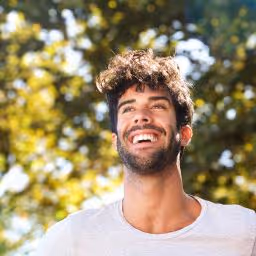 Smiling young man with dark curly hair and beard wearing a white T-shirt, outdoors with green trees in the background.