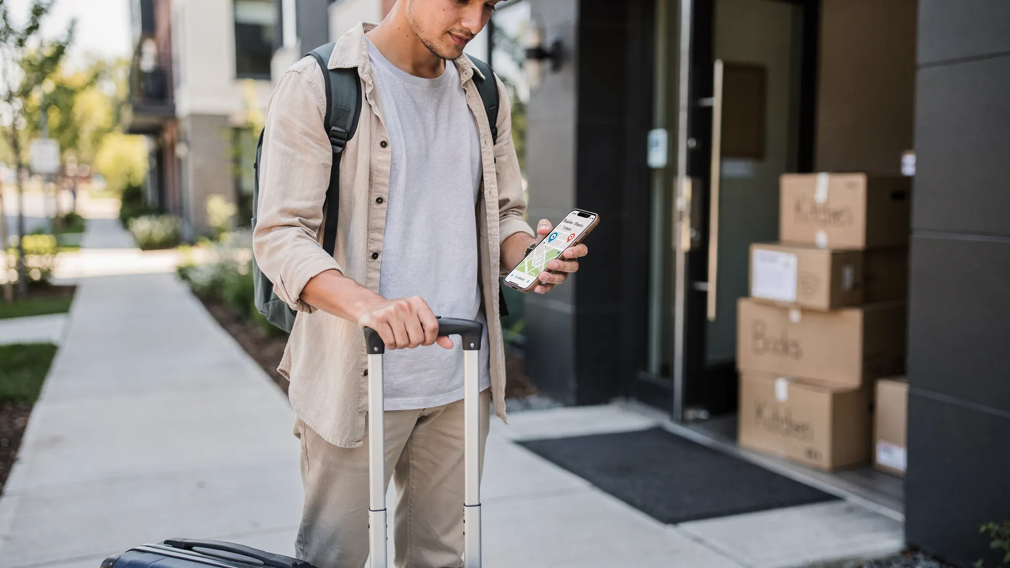 A person standing in a new city with a rolling suitcase, holding a phone showing a simple calendar and a neighborhood map (screen facing the viewer correctly), with moving boxes stacked in the background near a doorway, conveying a time-boxed short-term stay leading into a permanent rental.
