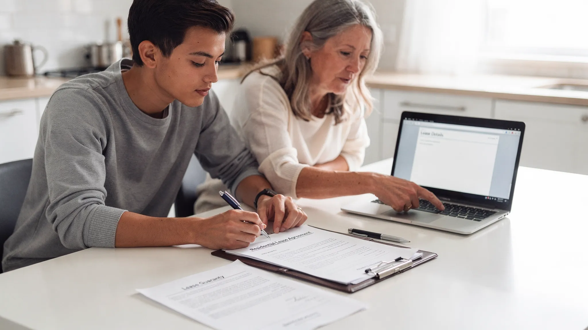 A renter and a supportive family member sitting at a kitchen table reviewing a residential lease and a separate guaranty form, with a pen, folder of documents, and a laptop facing the right direction.