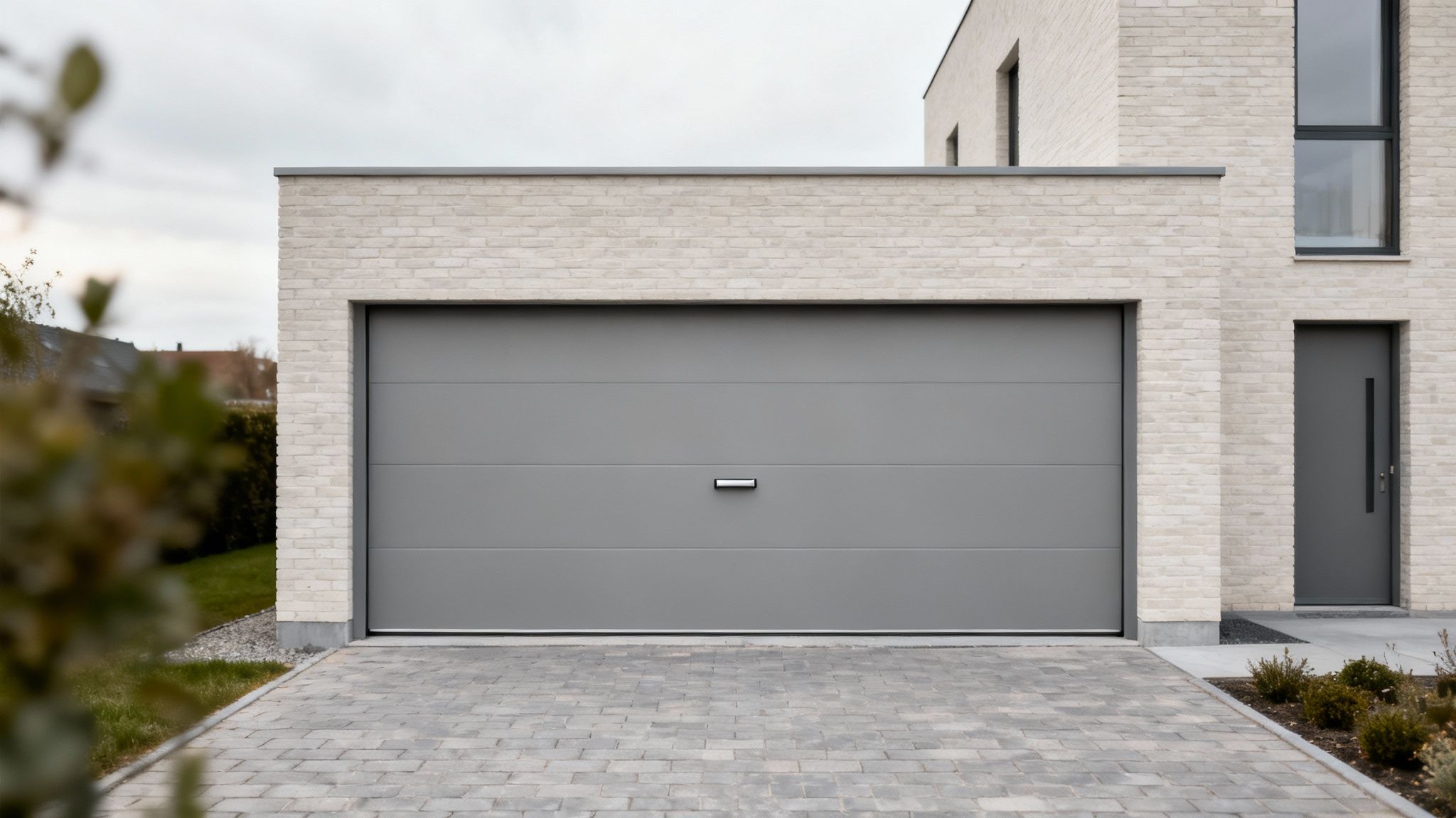 Maison moderne avec porte de garage sectionnelle grise et porte d'entrée assortie, façade en briques claires et allée pavée.