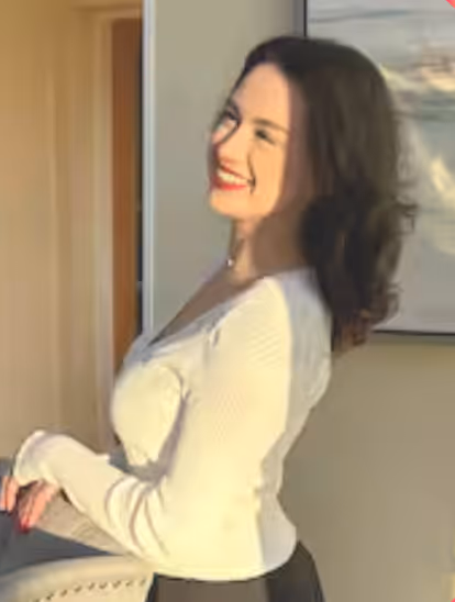 Smiling woman with dark hair wearing a white long-sleeve top, standing indoors near a chair and a framed picture.