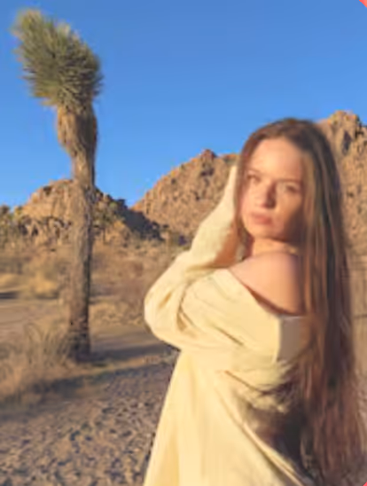 Young woman with long hair wearing a light-colored off-shoulder top, standing on a desert trail with rocky hills and a tall cactus under a clear blue sky.