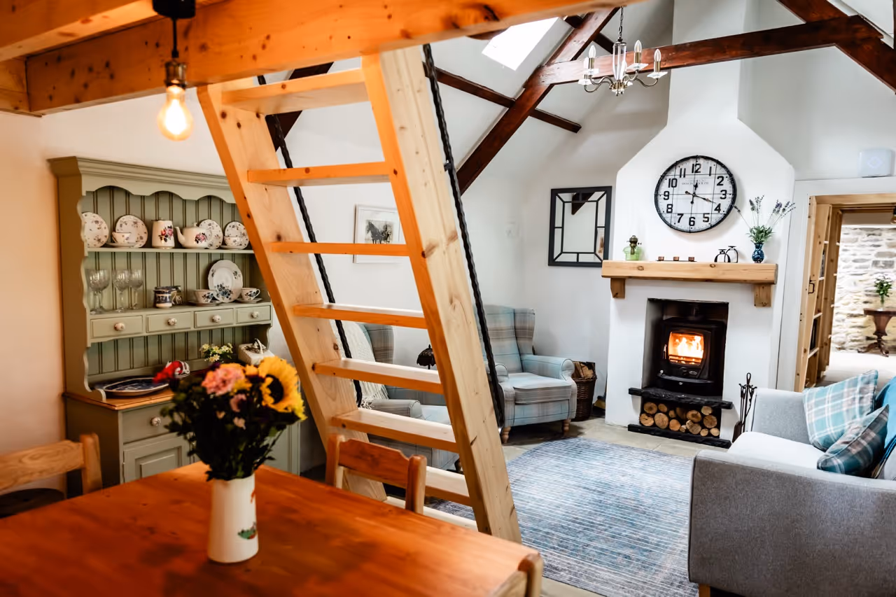 Cozy living room with wooden beams, ladder, armchairs, fireplace, clock, and a dining table with flowers.