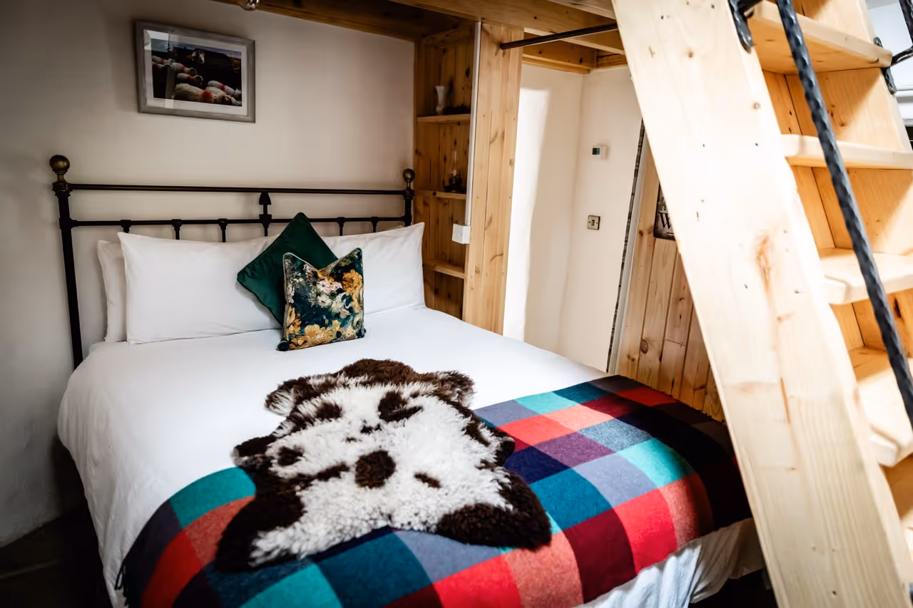 Cozy bedroom with white bed, green floral pillows, colorful plaid blanket, and wooden loft ladder.