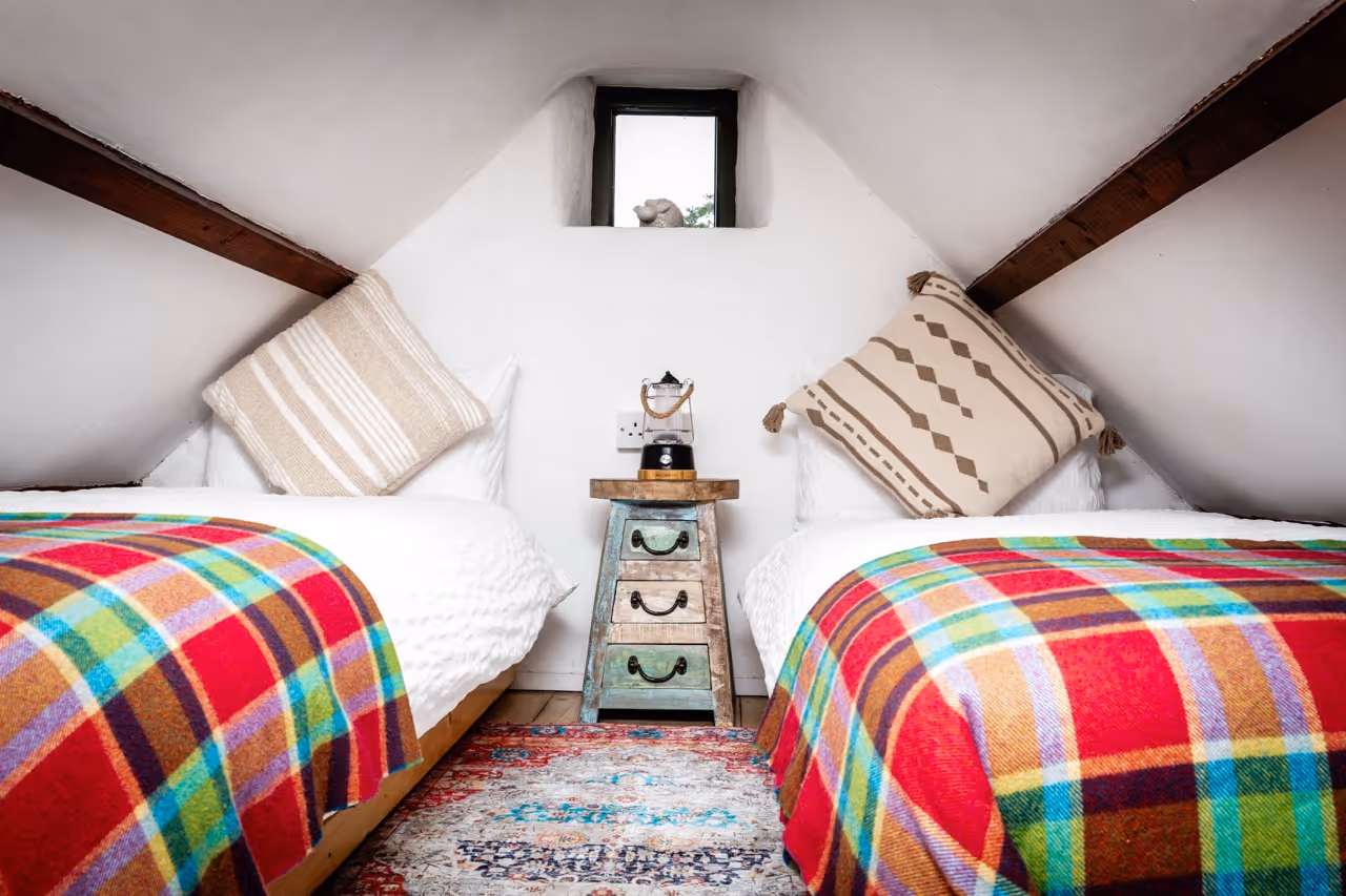 Attic bedroom with twin beds, colorful plaid blankets, patterned pillows, and a small rustic nightstand with a lantern.