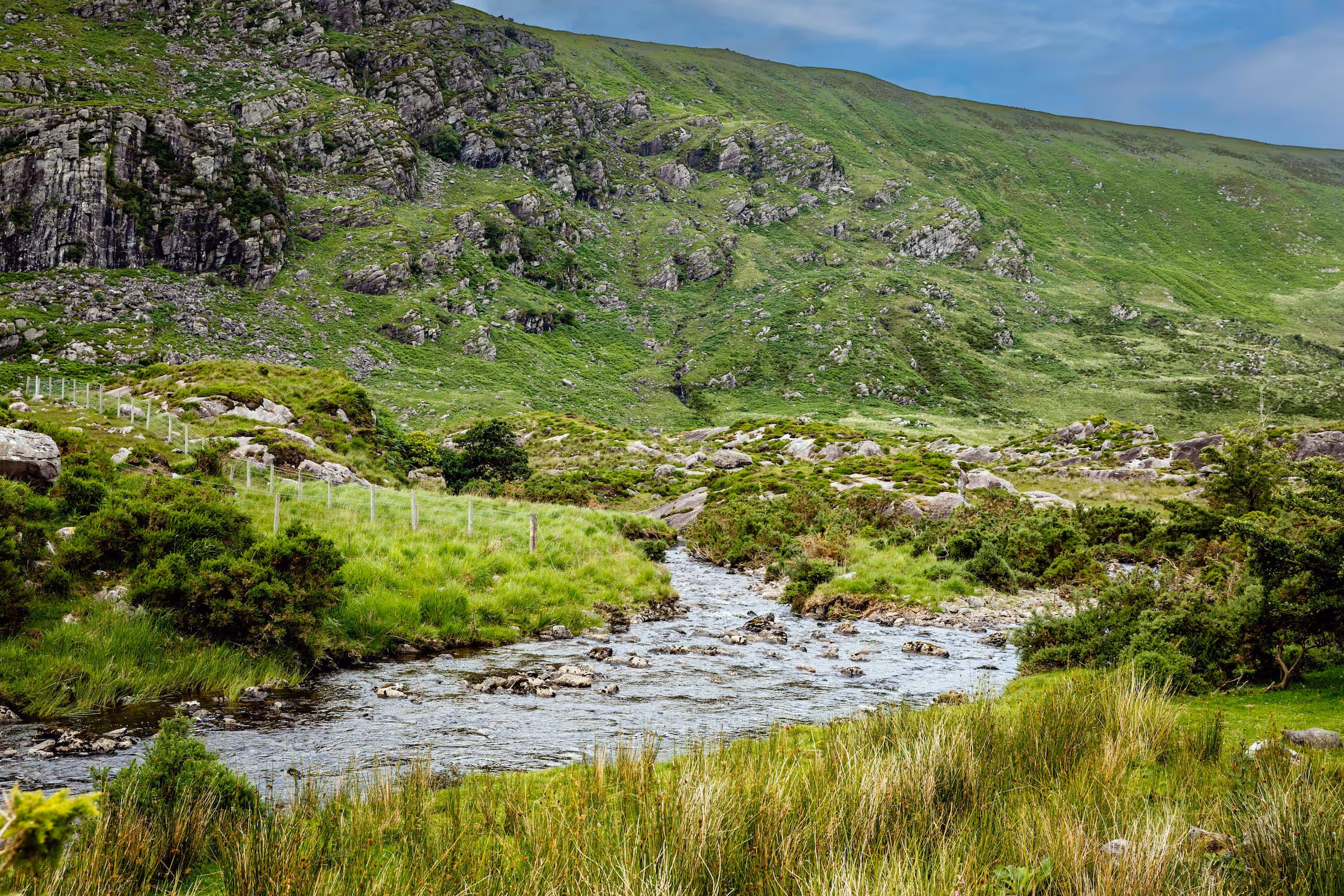 Small rocky stream flowing through lush green grass and shrubs at the base of a green mountain with rocky patches under a partly cloudy sky.