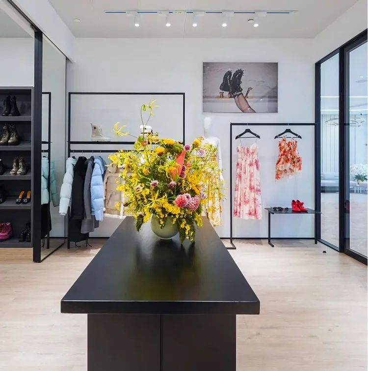 Chelsey House Showroom interior with black table featuring a large vase of colourful flowers, racks with coats and floral dresses, and shoes displayed on shelves.