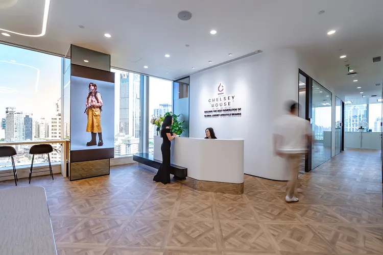 Chelsey House office reception area with a woman sitting behind a white desk talking to another woman, a man walking in the hallway, large windows showing cityscape, and a vertical digital screen displaying a woman in yellow pants.