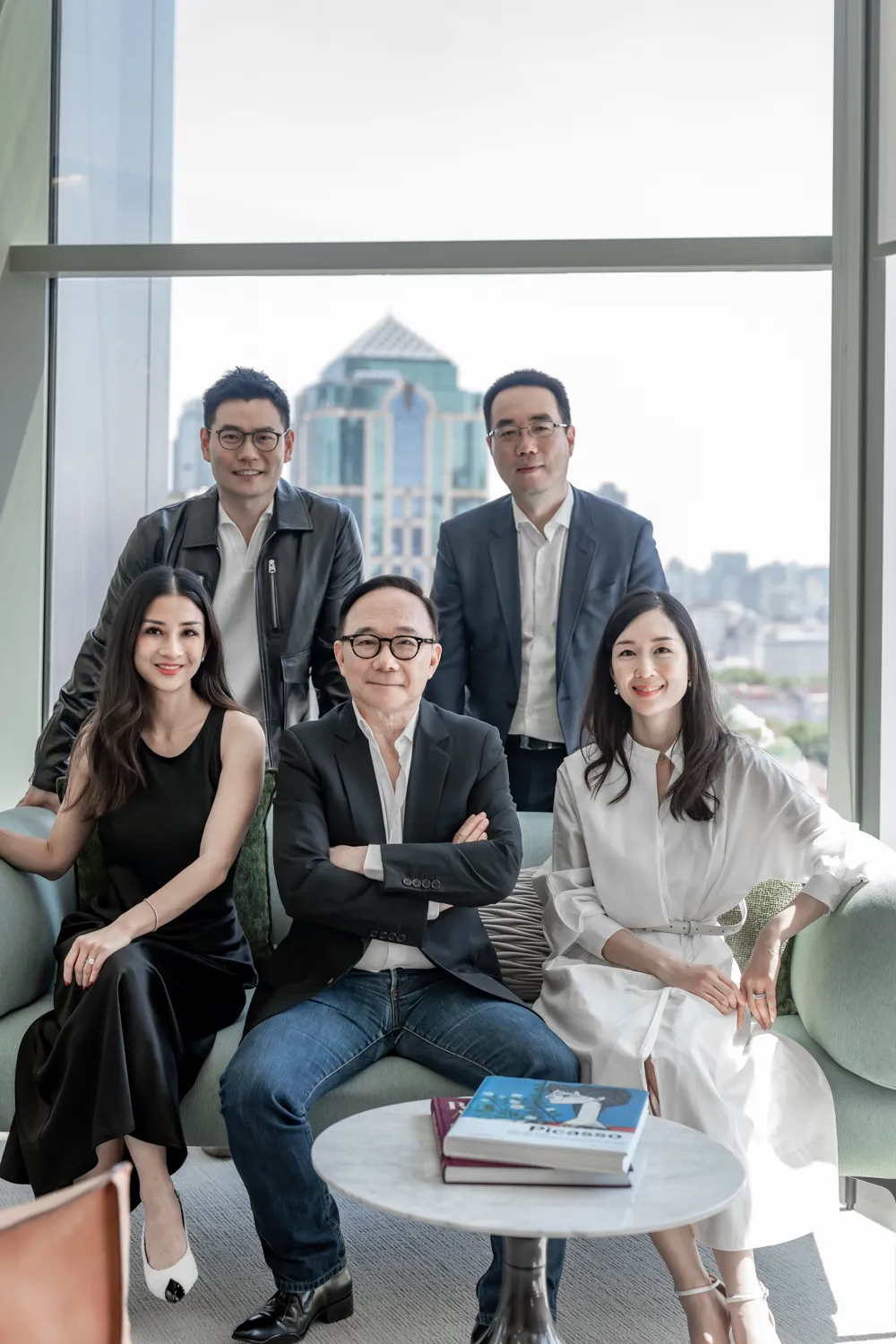 Group portrait of the Chelsey House core team. Three seated on a sofa and two standing behind, in a modern office with a city view through large windows.