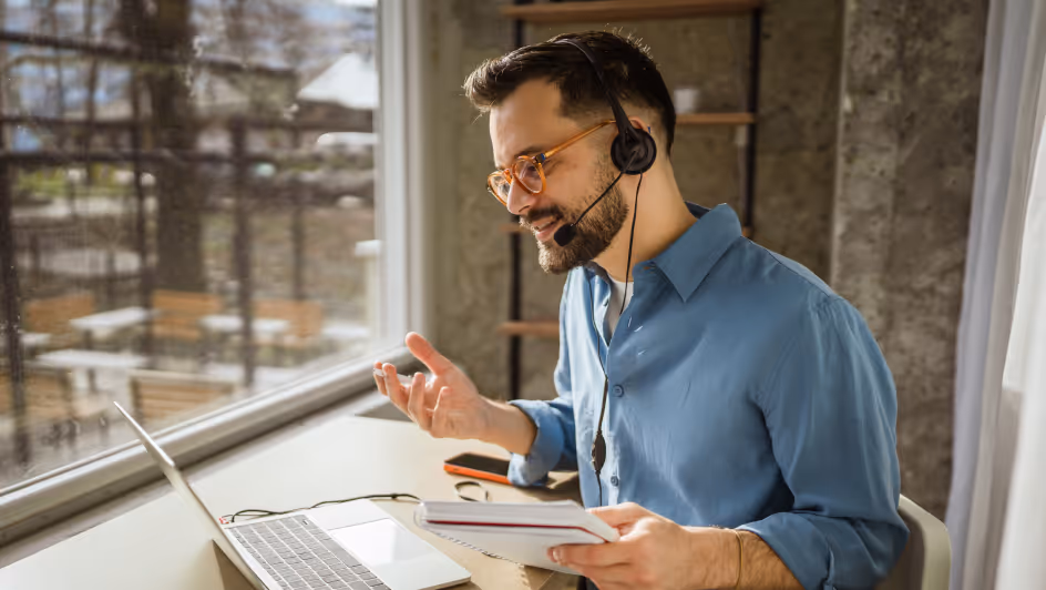 Man wearing glasses and a headset, sitting by a window and gesturing while looking at a laptop during a video call.