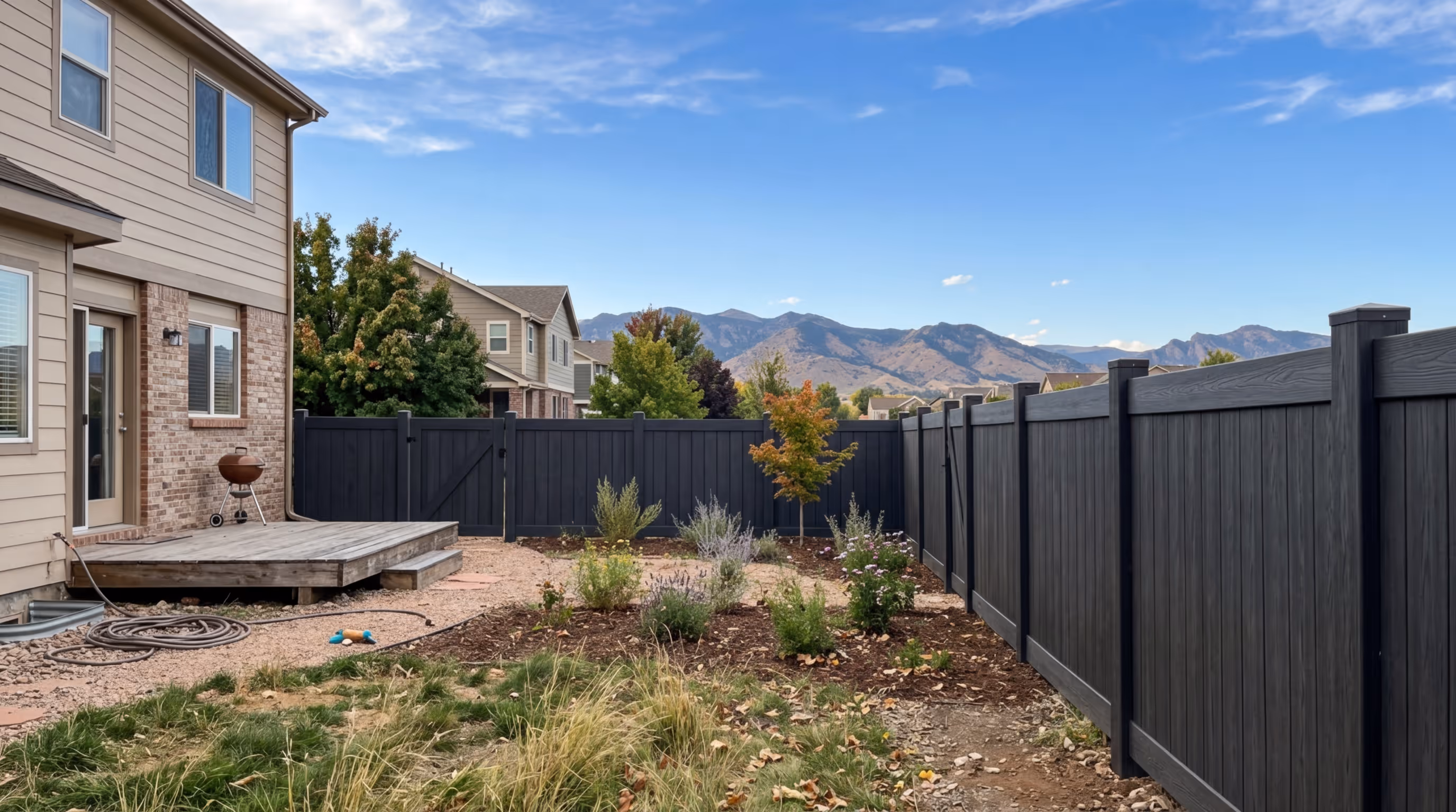 Backyard garden with new plants, black wooden fence, patio deck, and mountains in the background under a blue sky.