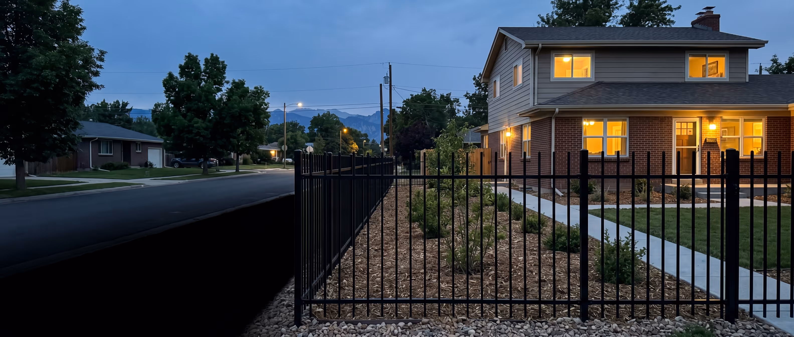 Two-story house with brick and gray siding, lit warmly at dusk, with a black metal fence and landscaped yard in front.
