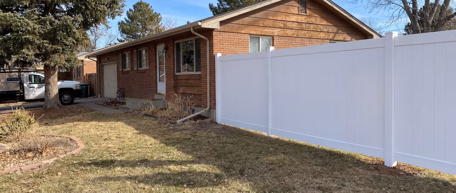 Front yard of a single-story brick house with a white vinyl fence, a large tree, and a parked white truck.