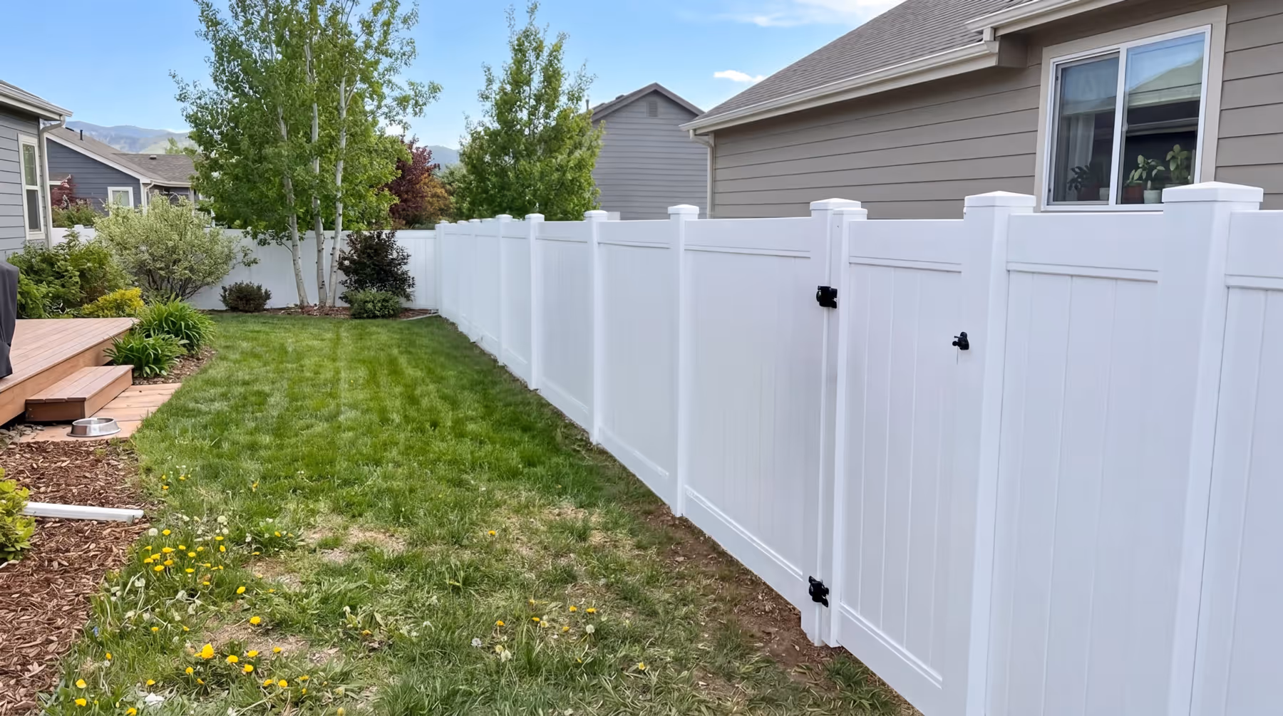 Backyard with green grass, wooden deck, bushes, and a long white vinyl privacy fence with a gate.