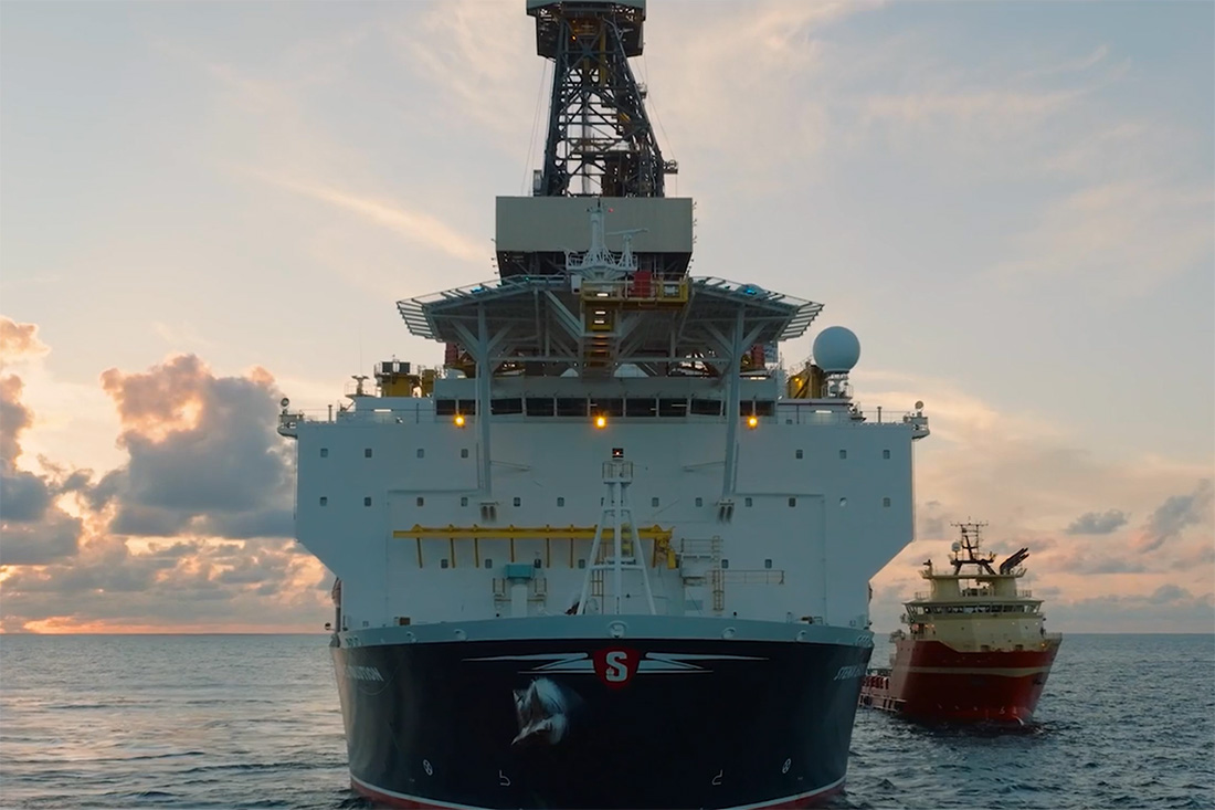 Front view of a large offshore drilling ship with a smaller support vessel behind it on the ocean at sunset under partly cloudy sky.