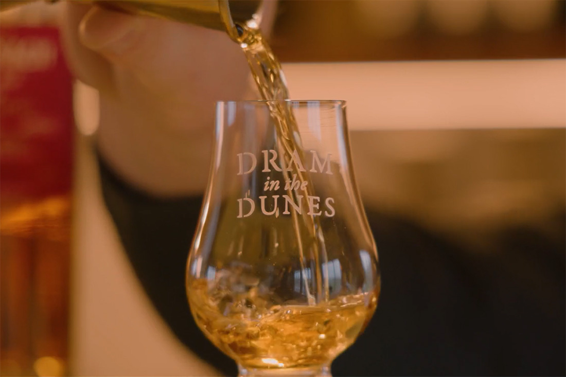 A golden-brown liquid is being poured into a clear glass with the words 'DRAM in the DUNES' etched on it.